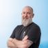 A man with a beard standing in front of a blue background.